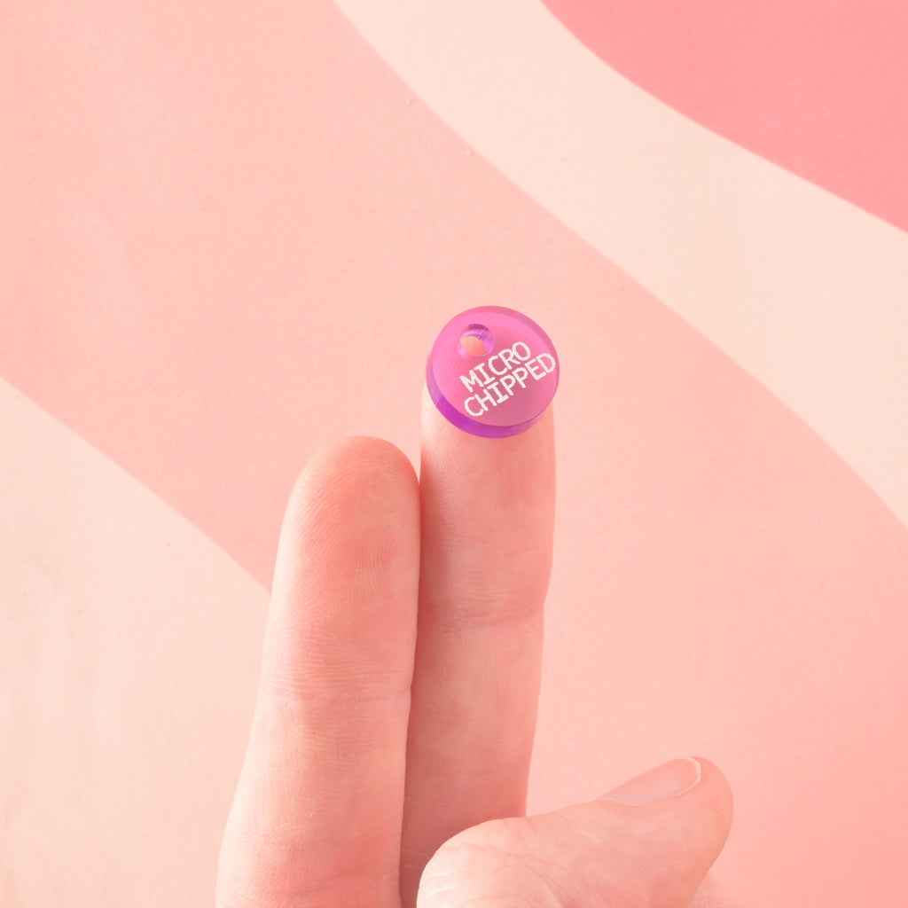 A small, circular acrylic charm in translucent purple engraved with "MICROCHIPPED" in white text. The tiny charm is held up on the tip of a finger to show scale.