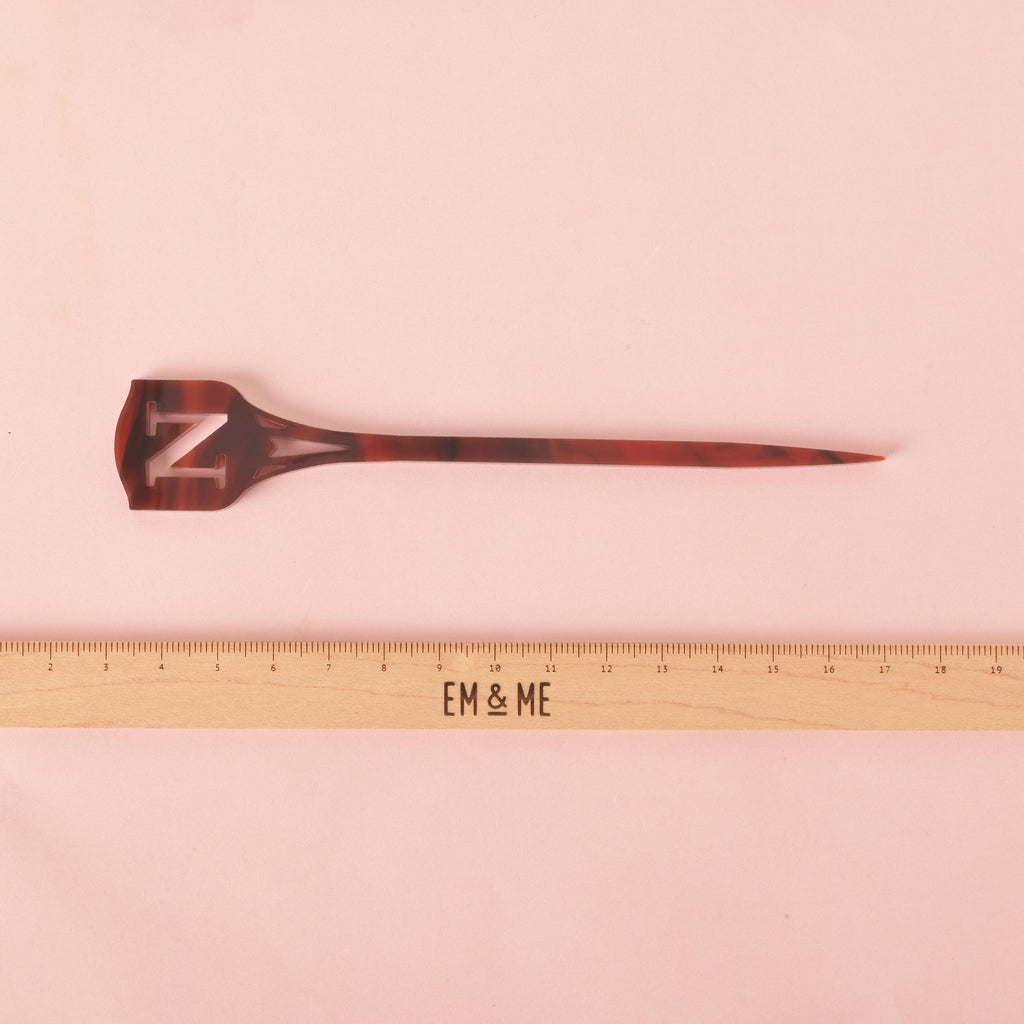 A brown and black tortoiseshell cocktail stirrer featuring the cutout monogram letter 'N' at the top. The stirrer has a pointed shield shape. A wooden ruler with the "EM & ME" logo is placed below the stirrer to show its size. The item is displayed on a solid pink background.