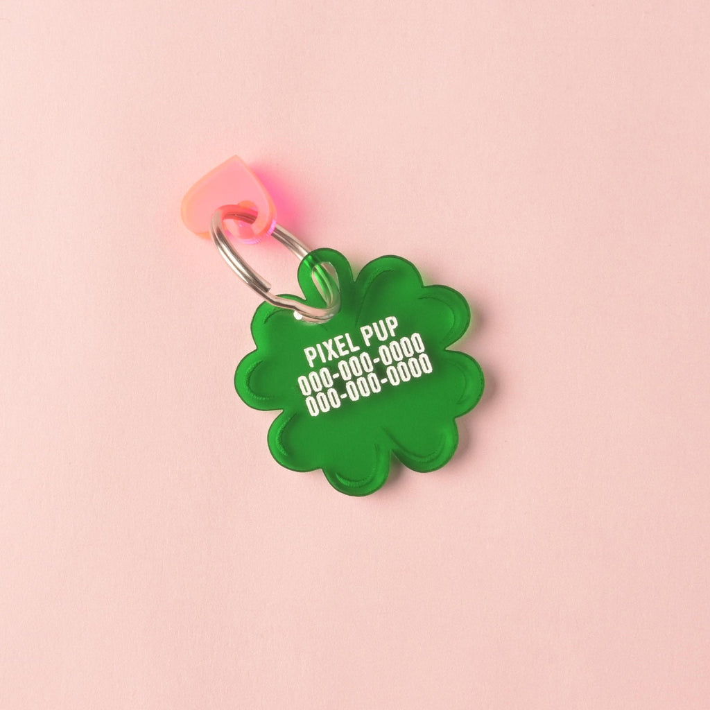 Close-up of a translucent green, clover-shaped acrylic pet ID tag. The tag is personalized with the name 'PIXEL PUP' and placeholder phone numbers in white text. A translucent pink heart charm is attached to the silver split ring, all against a solid pink background. Lucky AF, 4 leaf Clover Personalized Pet Tag, St Patrick Cat and Dog ID Tag