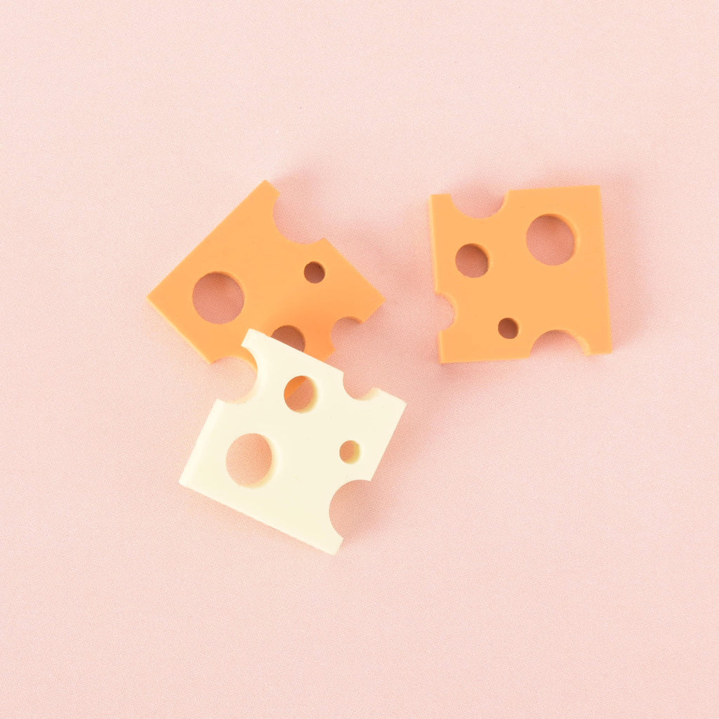 A group of three cheese-shaped charms. One is a light yellow-white color, while the other two are a more vibrant orange-yellow. The charms have various-sized holes and are shown on a light pink background.