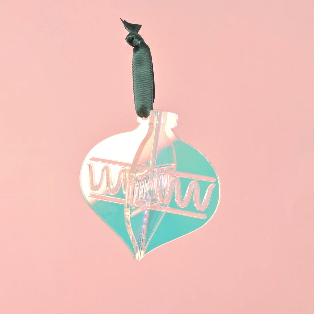 A single 3D interlocking acrylic Christmas ornament in a stylized bulb shape, featuring iridescent and translucent teal pieces. The ornament has engraved zigzag patterns and hangs from a dark green ribbon against a light pink background. Playful Christmas ornaments, bright colors and non-breakable, kid Christmas