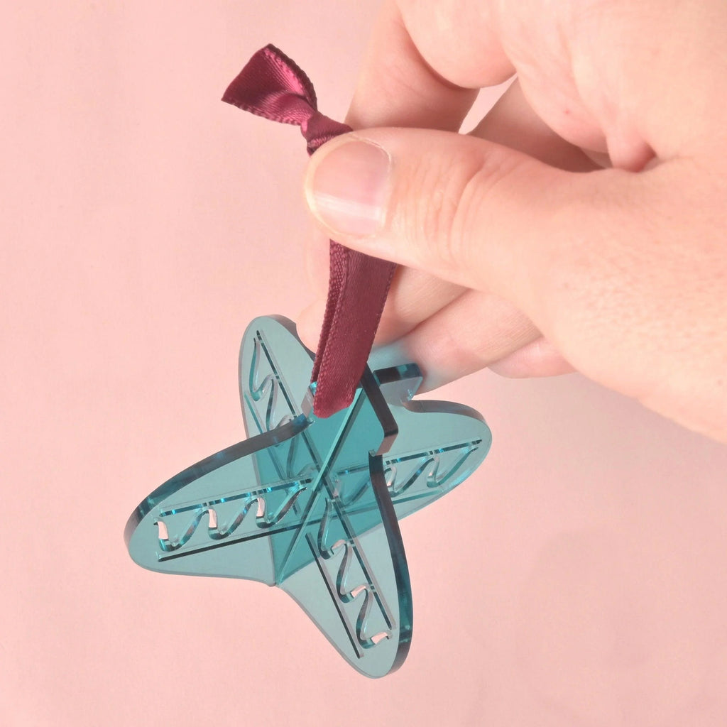 A hand holds a 3D interlocking acrylic Christmas ornament in a stylized, abstract star shape. The ornament is translucent teal/blue-green and features engraved zigzag patterns. It hangs from a dark red ribbon against a light pink background. Playful Christmas ornaments, bright colors and non-breakable, kid Christmas