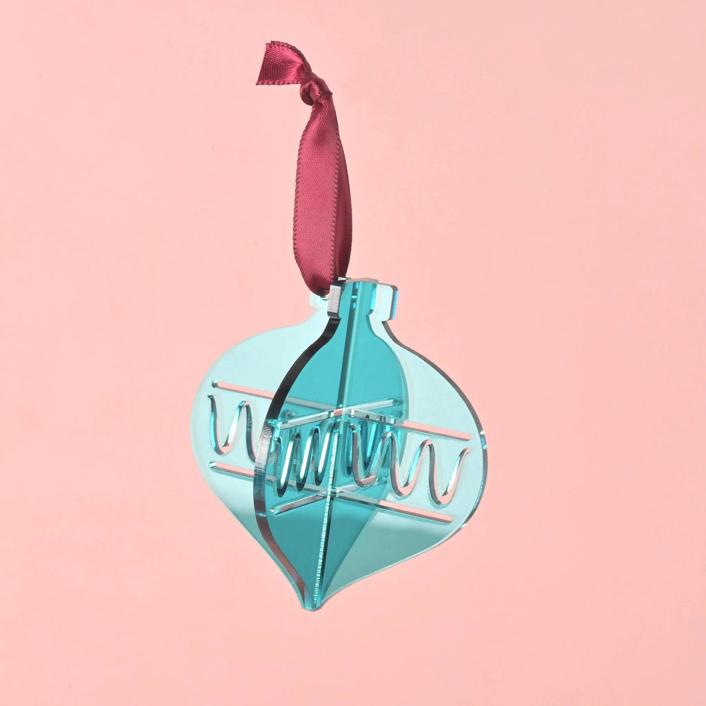 A single 3D interlocking acrylic Christmas ornament in a translucent blue-green (teal) color, featuring an engraved zigzag pattern on the inner layer. The ornament hangs from a dark red ribbon against a solid pink background. Playful Christmas ornaments, bright colors and non-breakable, kid Christmas