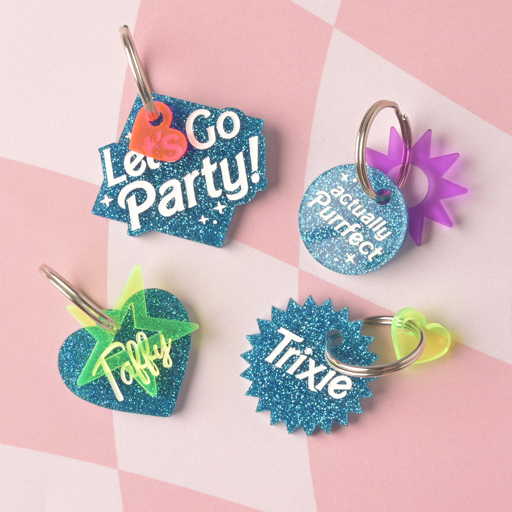 A collection of four blue glitter acrylic tags: one with the slogan "Let's Go Party!" and an orange heart charm; one round tag with "actually Purrfect" and a purple sunburst charm; a heart tag for "Taffy" with a neon star accent; and a starburst tag for "Trixie" with a neon heart charm.