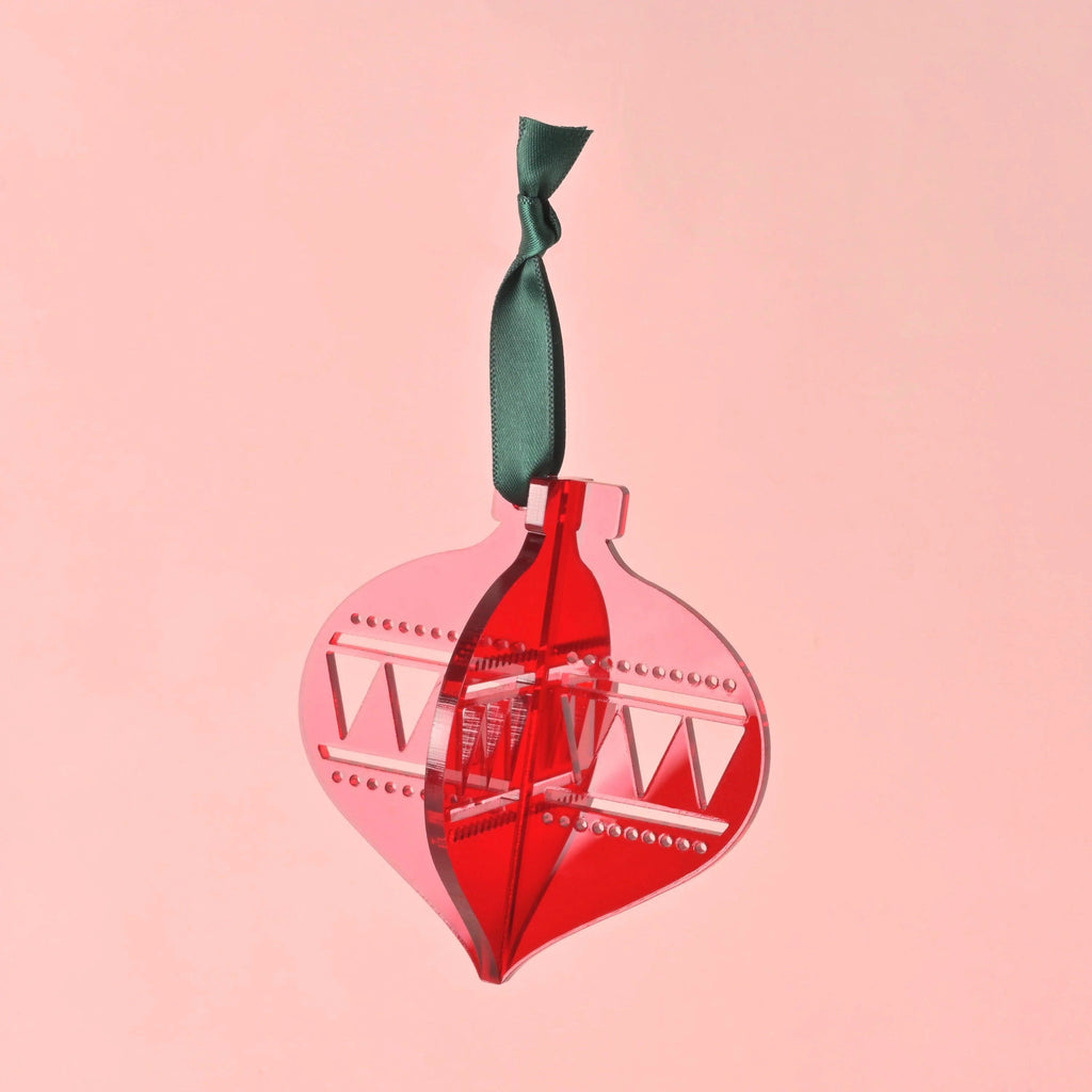 A single 3D interlocking acrylic Christmas ornament in layered red acrylic, featuring cut-out geometric and triangle patterns. The ornament hangs from a dark green ribbon against a solid pink background.Acrylic Christmas ornaments, bright colors and non-breakable