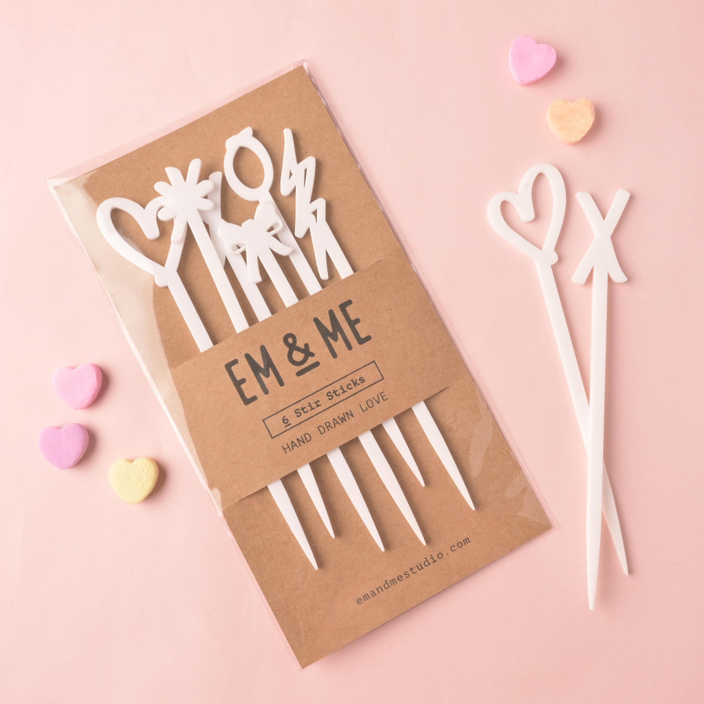 A packaged set of EM & ME white acrylic 'Hand Drawn Love' stir sticks (6 pieces) in a clear sleeve with a kraft paper backing. Two loose white stir sticks (a heart and an XO shape) and scattered pastel conversation heart candies are displayed nearby on a pink background.