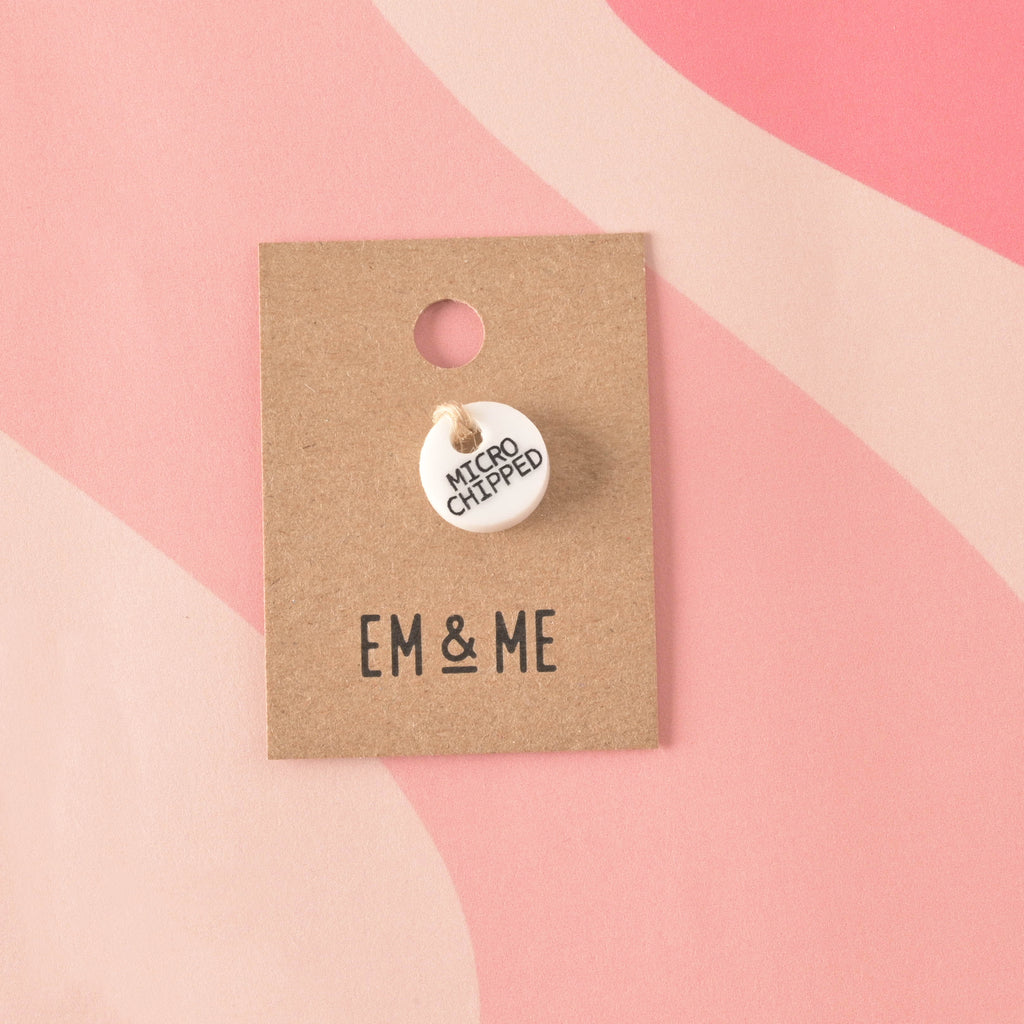 A small, circular, opaque white acrylic charm engraved with "MICROCHIPPED" in black text. The charm is secured with twine to a branded 'EM & ME' kraft card.