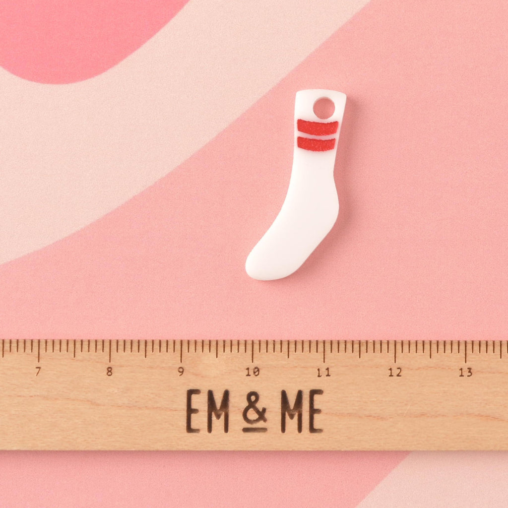 Tubesock, Missing Sock, Seasonal Mini, Tiny Accessory for Pet ID  A small, opaque white acrylic mini charm shaped like a tube sock with two red stripes near the top. The charm is shown above a wooden ruler with the 'EM & ME' logo to indicate its size.Tag, Cats and Dogs Add-on Charm