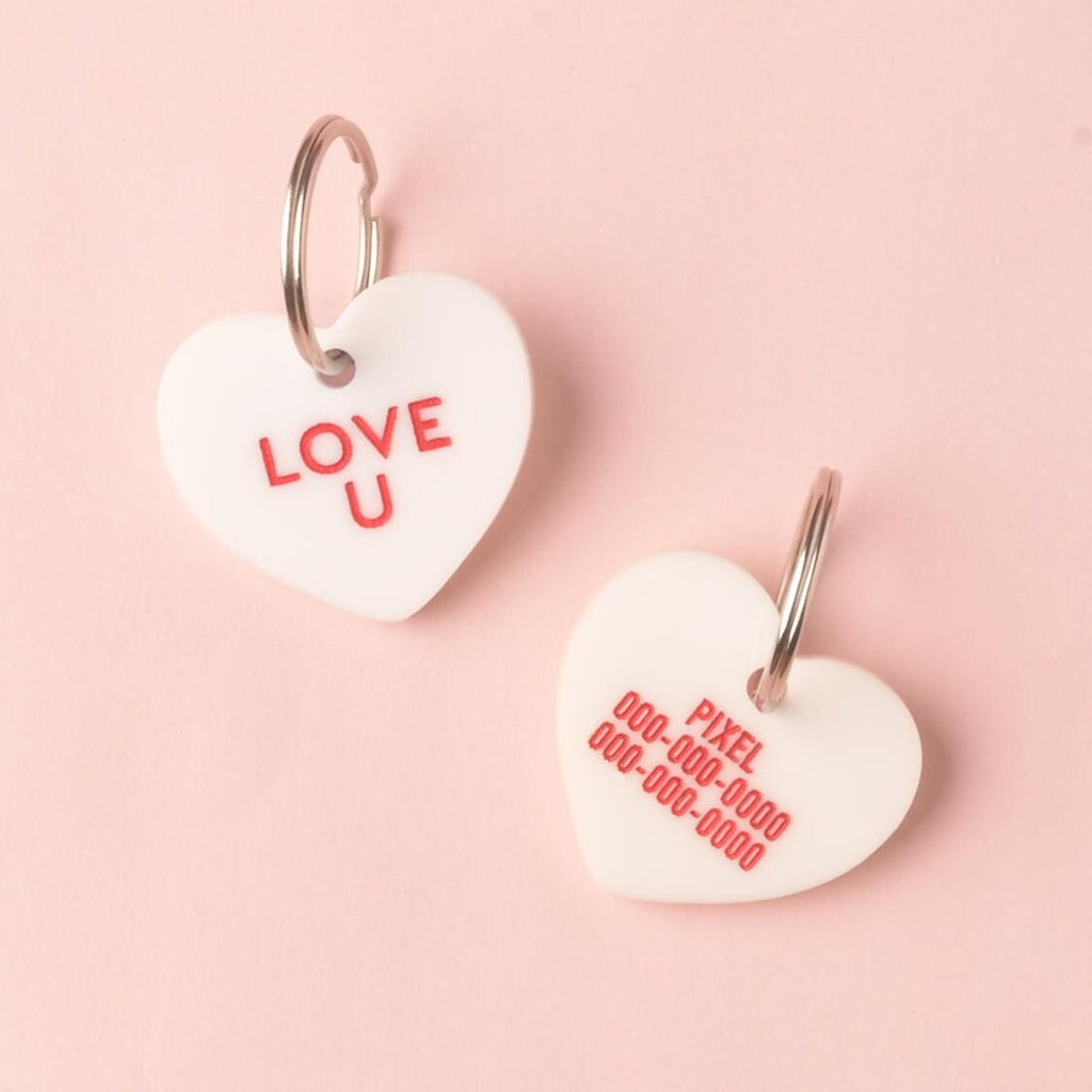 Two views of a white heart-shaped acrylic pet ID tag on a pink background. One side shows the red conversation heart slogan 'LOVE U.' The other side shows personalized safety details for 'PIXEL' with red placeholder phone numbers. Conversation Hearts, Valentines Heart Dog tag, Customize front and back, ID tag for cats and dogs, Personalized Pet Tag