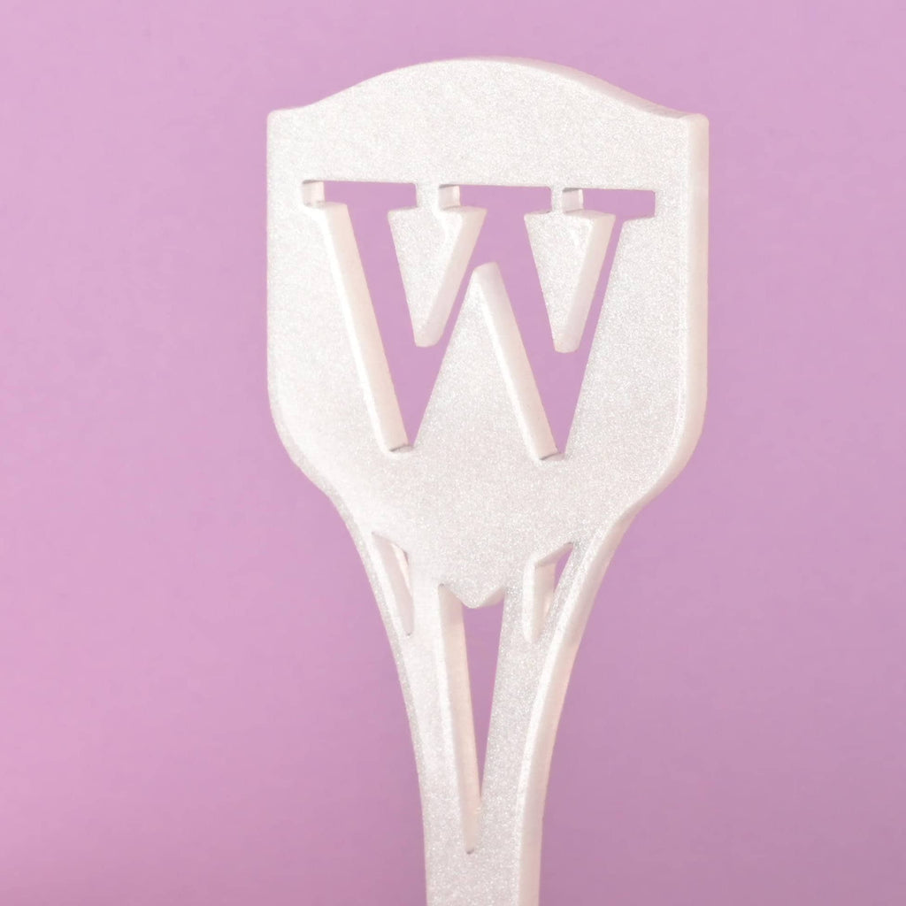 A close-up of a white iridescent cocktail stirrer featuring the cutout monogram letter 'W' at the top. The stirrer has a pointed shield shape and is viewed against a solid pink background.