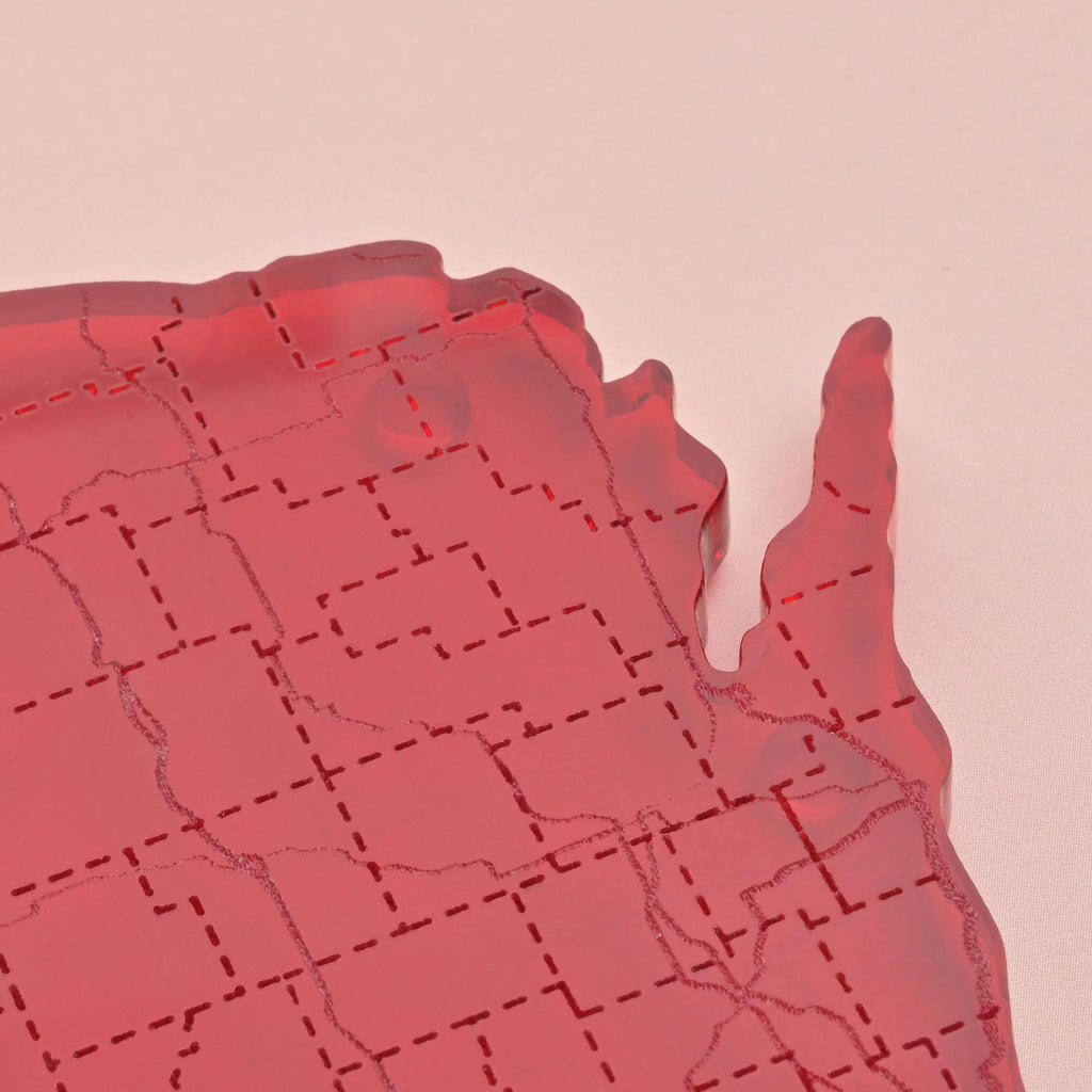 Close-up of the upper right corner of a red acrylic Wisconsin-shaped coaster showing fine etched map lines and smooth laser-cut edges on a soft pink background.