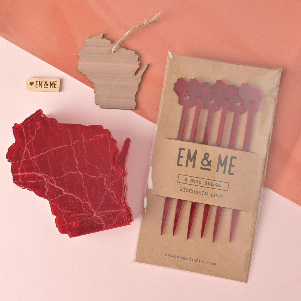Wisconsin-themed flat lay featuring a pack of red acrylic drink stir sticks, a red acrylic state map coaster, a wooden Wisconsin ornament with twine hanger, and a small engraved EM & ME Studio logo tag, all arranged on a peach and pink background.