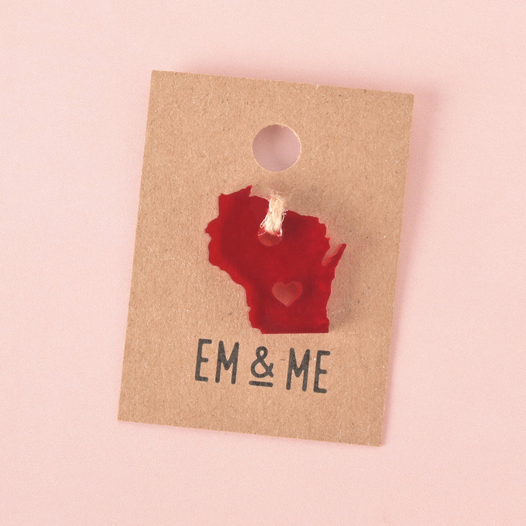 Red acrylic Wisconsin state charm with heart cutout, tied with twine and displayed on a kraft paper card featuring EM & ME Studio branding, set against a soft pink background.
