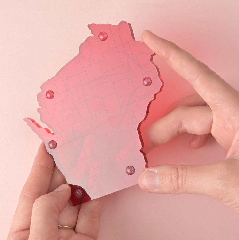 Close-up of a red acrylic coaster shaped like Wisconsin, featuring etched map details and five clear rubber feet, held between two hands on a soft pink background.