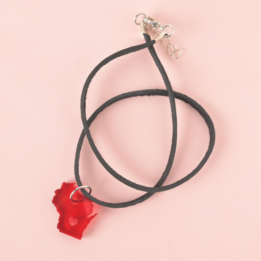 Red acrylic Wisconsin state-shaped pendant with heart cutout, hanging from a black faux suede cord necklace with silver clasp, displayed on a soft pink background.