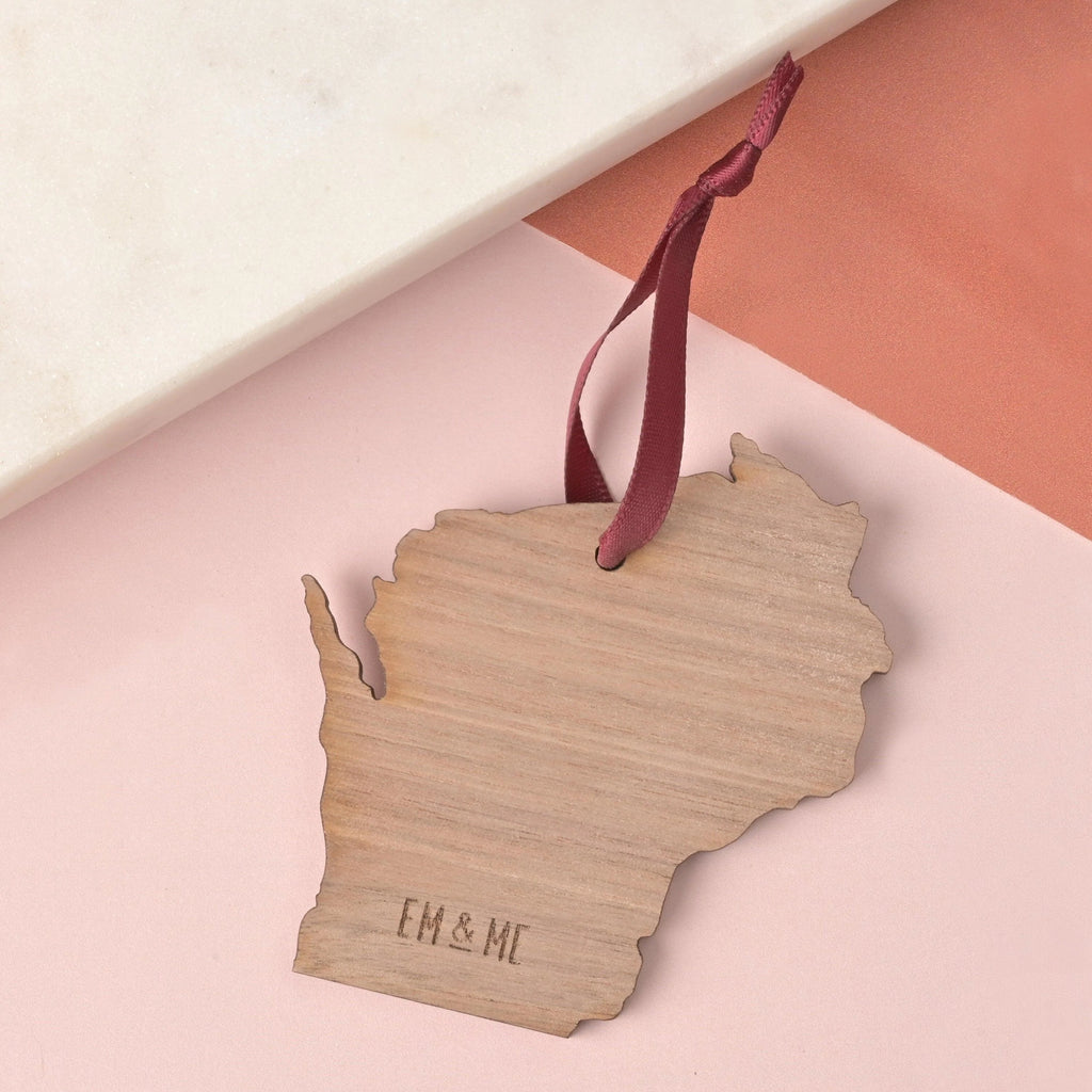 Wisconsin-shaped wooden ornament with laser engraved EM & ME logo and burgundy ribbon hanger, displayed on a peach and blush-toned background with marble accent.