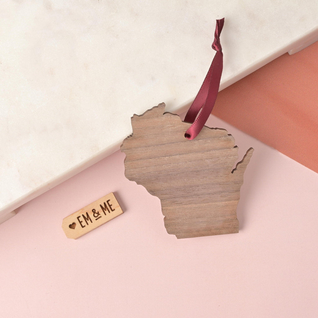 Wooden Wisconsin state ornament with a burgundy ribbon hanger, placed on a marble and blush pink background next to a small engraved EM & ME wooden tag.
