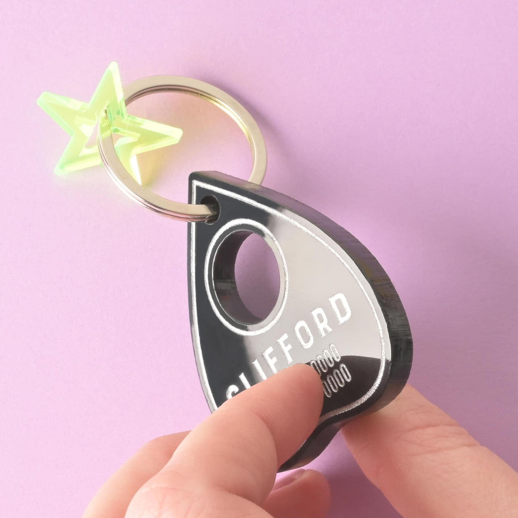 A person's hand holds a personalized black Ouija planchette-shaped pet tag. The tag has the name "CLIFFORD" and a placeholder phone number. A small, translucent green star charm is attached. The items are shown against a solid purple background.