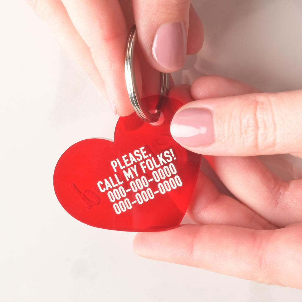 A person's hands hold a personalized red heart-shaped pet tag. The tag says "PLEASE. CALL MY FOLKS!" and has a placeholder phone number on it. The tag is attached to a silver ring, perfect for a collar, and is shown against a white background.