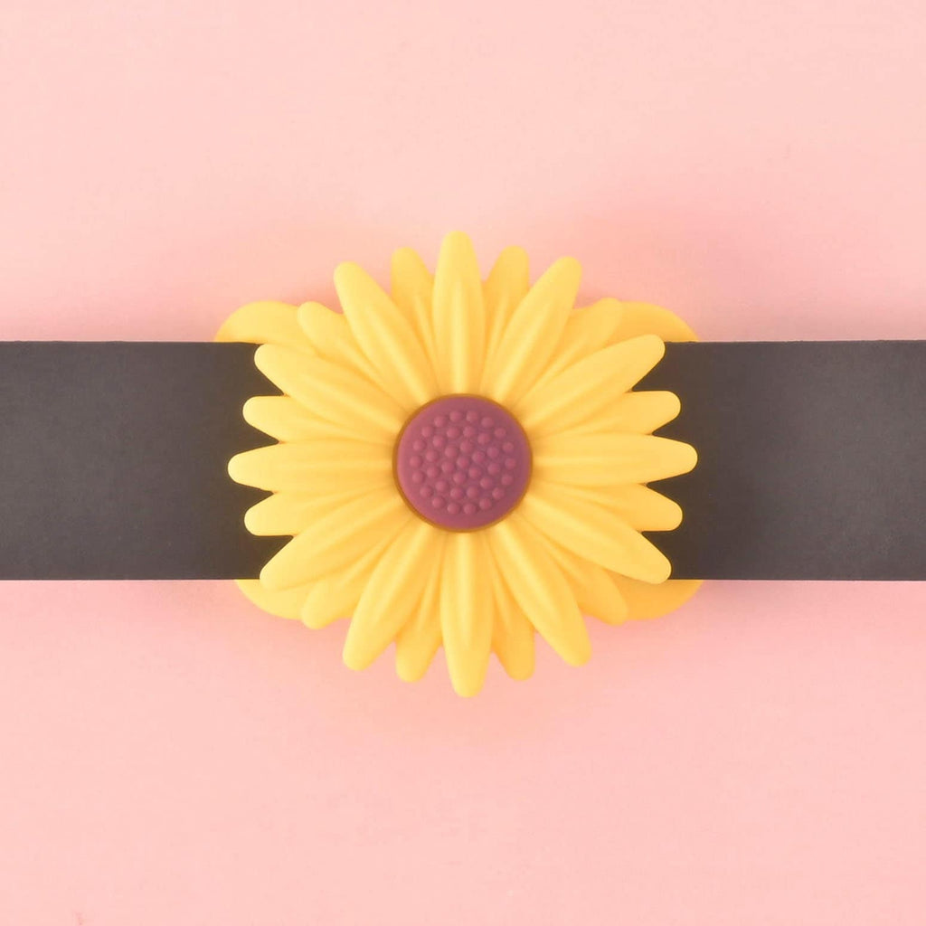 A yellow flower-shaped pet AirTag holder with a brown center, attached to a black collar strap. The holder is displayed on a solid pink background.
