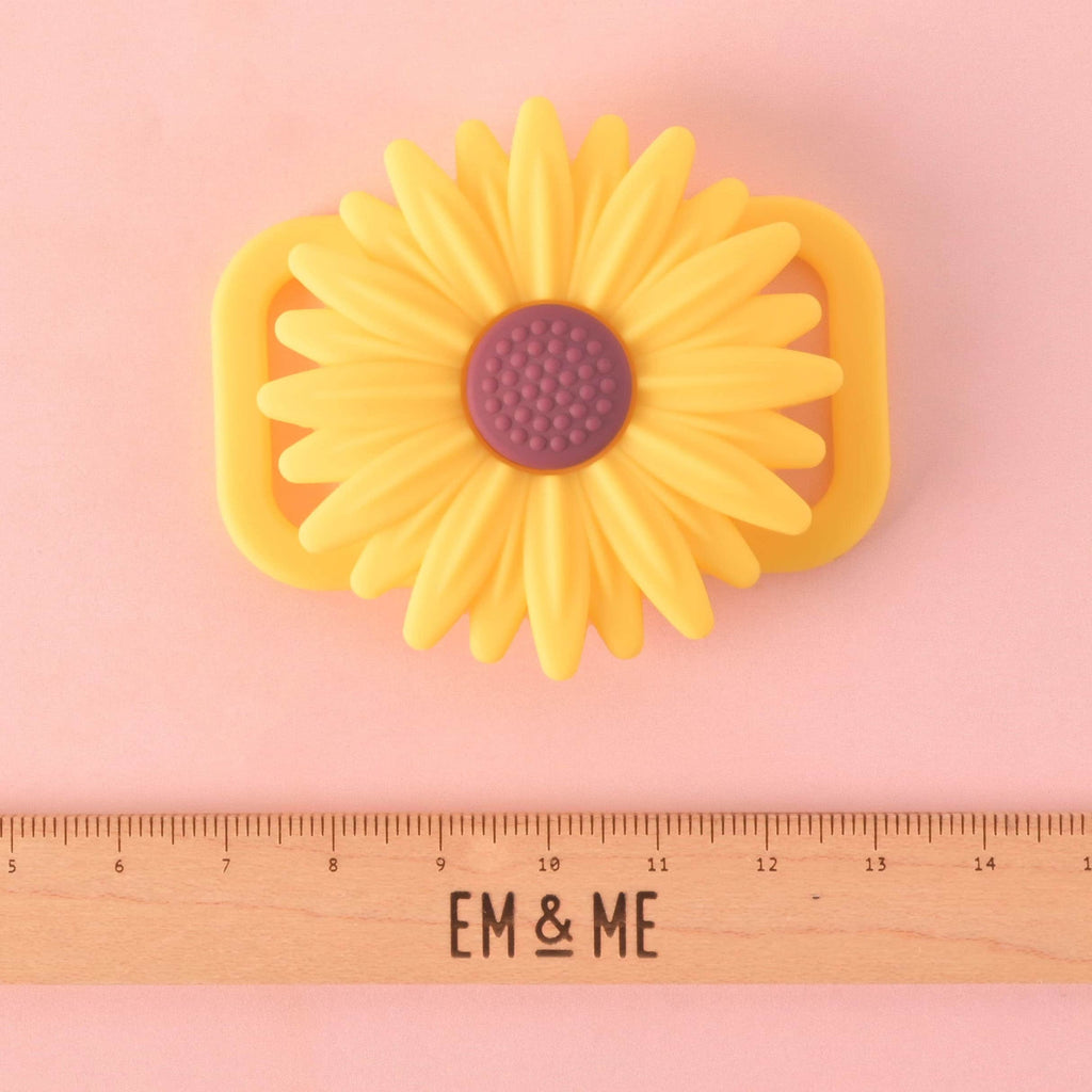 A yellow flower-shaped pet AirTag holder with a purple center. A wooden ruler with the "EM & ME" logo is placed below the holder to show its size. The holder is displayed on a solid pink background.