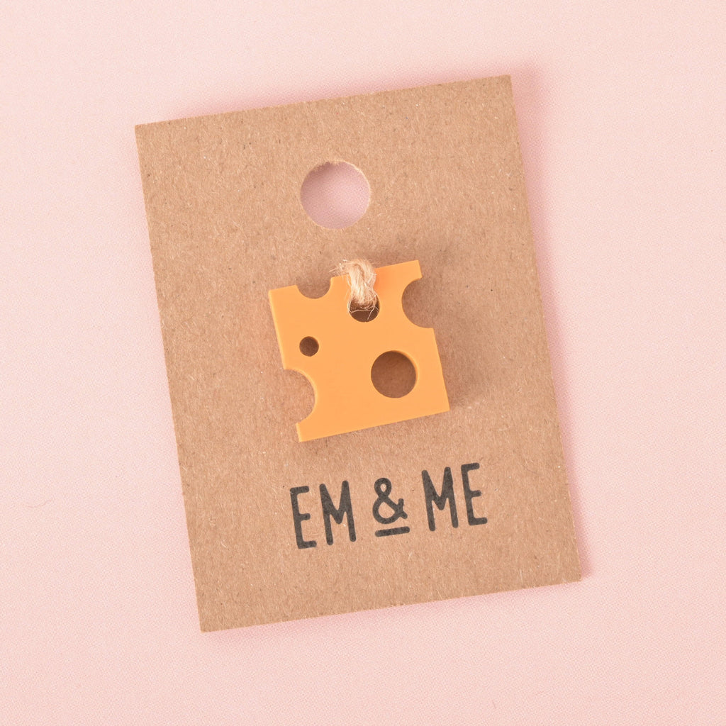 A single orange, cheese-shaped pin or charm attached to a brown paper card that reads "EM & ME." The item is displayed on a solid pink background.