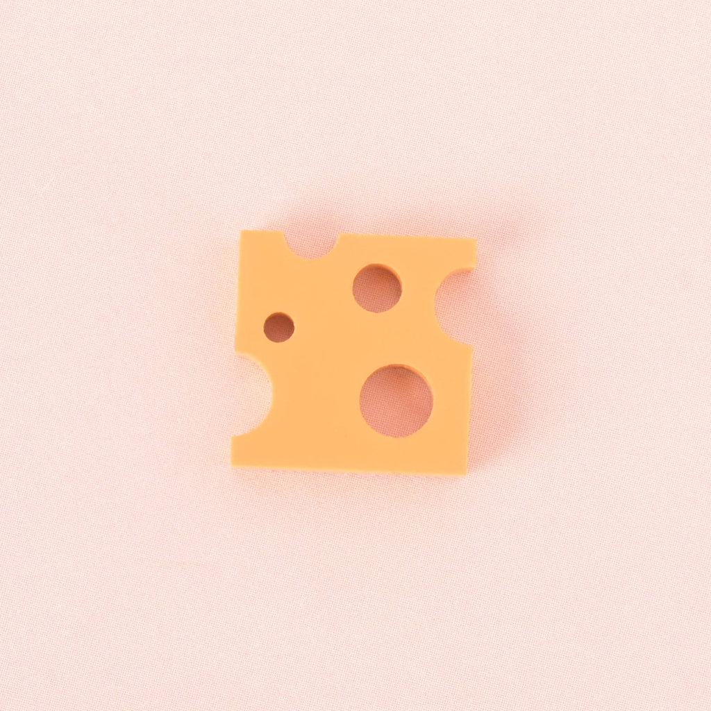 A single Yellow orange, cheese-shaped cheddar charm with various-sized holes. The item is shown on a solid pink background.