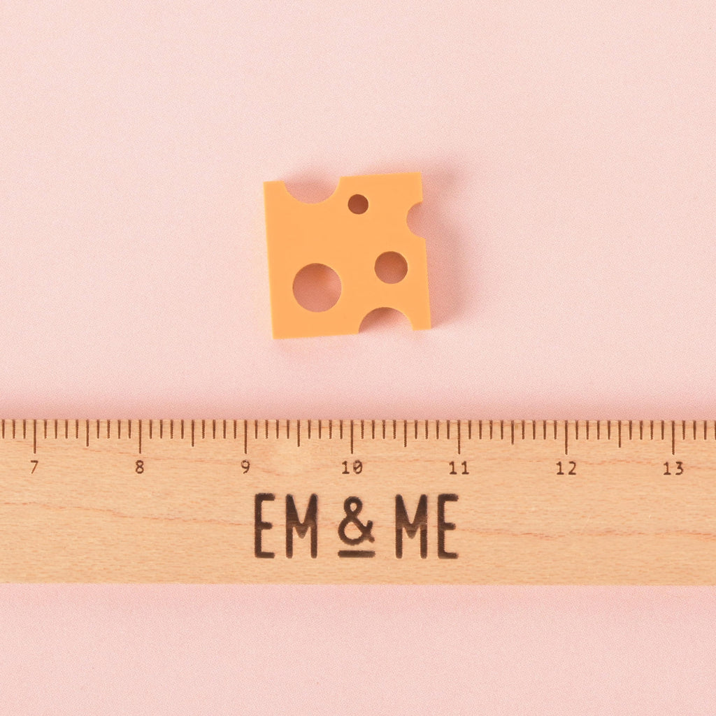 A single orange, cheese-shaped charm with various-sized holes. A wooden ruler with the "EM & ME" logo is placed below the charm to show its size. The charm is shown on a solid pink background.