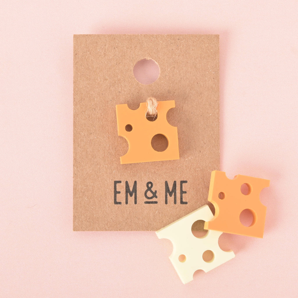 A single orange cheese-shaped charm is attached to a brown paper card that reads "EM & ME." Two other cheese charms, one white and one orange, are shown below the card. The items are displayed on a solid pink background.