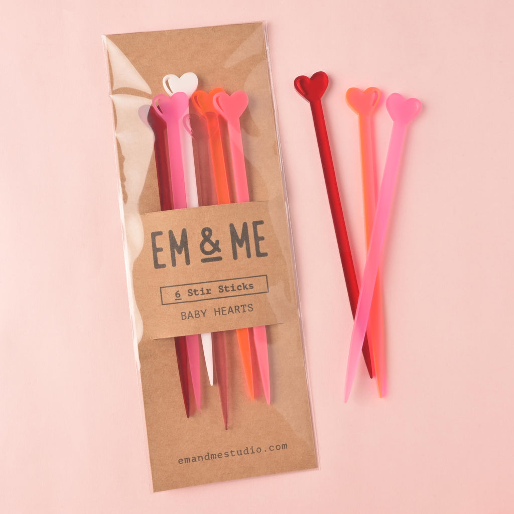 A packaged set of EM & ME 'Baby Hearts' acrylic stir sticks (6 pieces in pink, red, orange, and white) in a clear sleeve with a kraft paper backing. Three loose stir sticks in red and pink are lying beside the package, all on a light pink background.