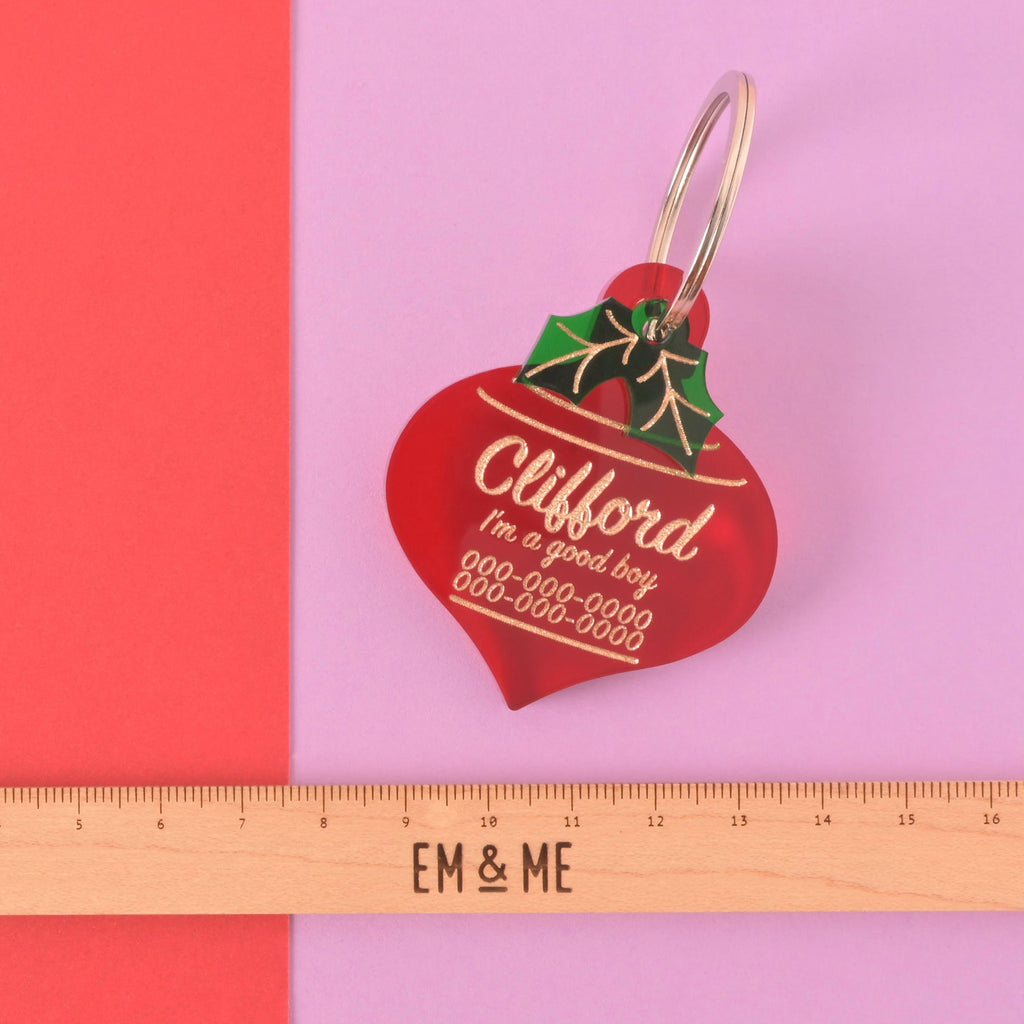 Clifford red ornament-shaped acrylic dog tag with holly detail shown above wooden EM & ME ruler for size reference on pink background