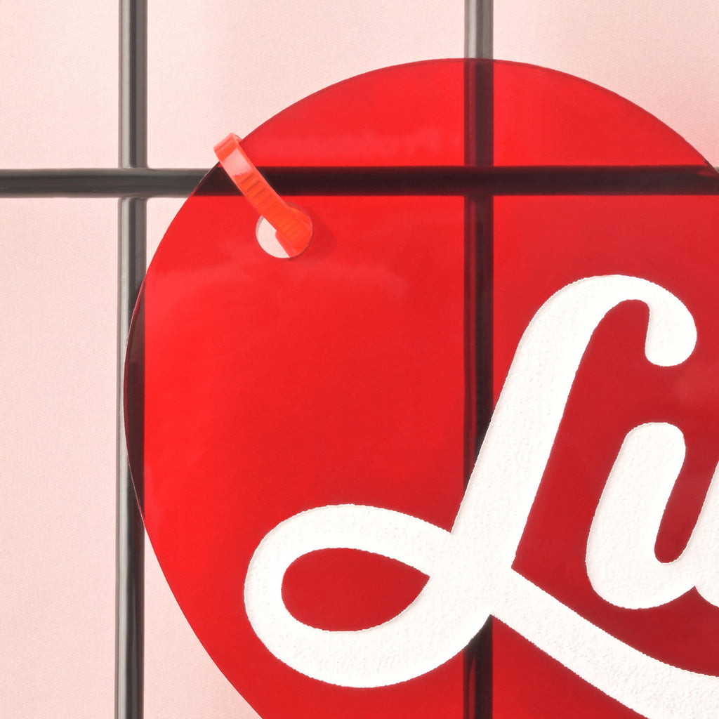 Close-up of red acrylic heart crate nameplate attached to a black wire crate with an orange zip tie; partial white script name “Lucy” visible.
