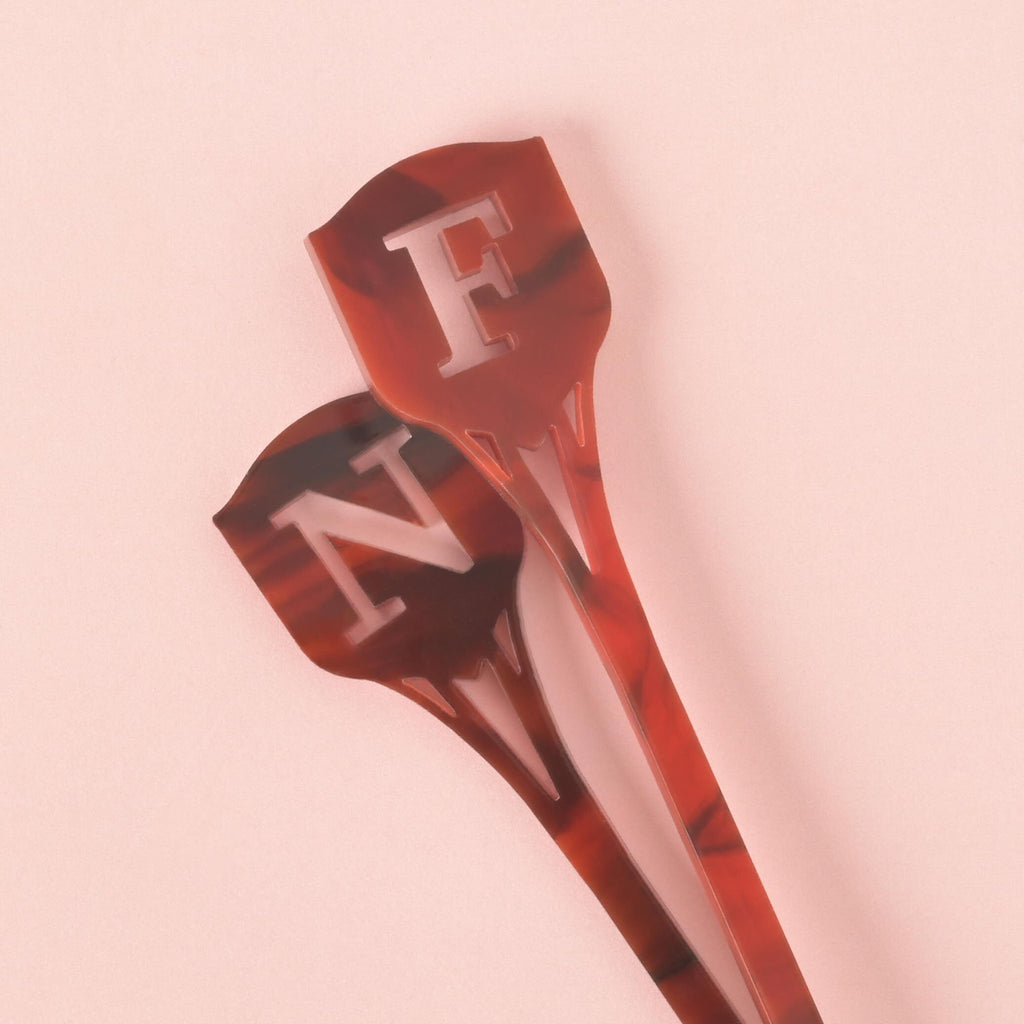 A close-up of two dark red and black tortoiseshell cocktail stirrers. The stirrers feature the cutout monogram letters 'F' and 'N' at the top in a shield shape, overlapping each other on a solid pink background.