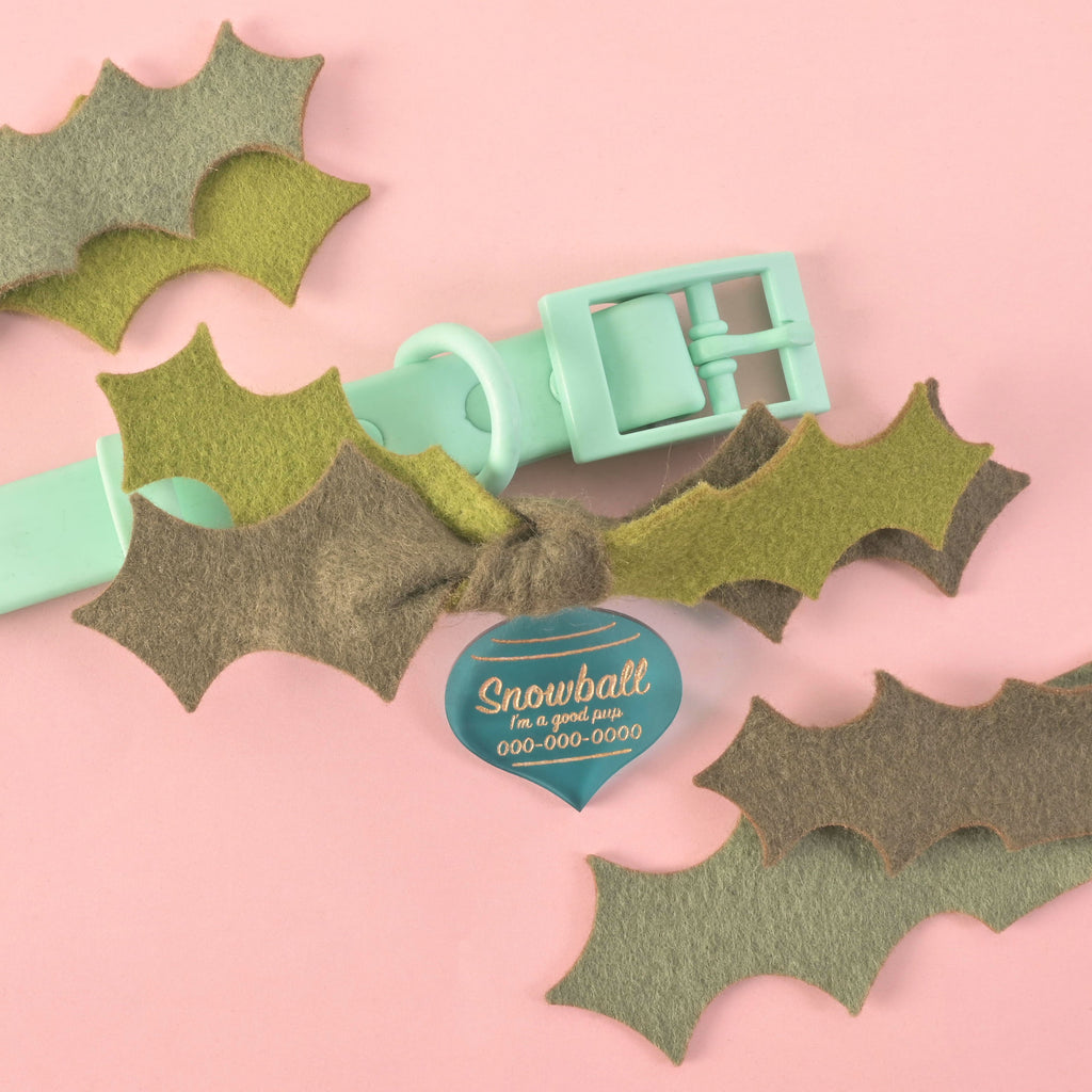 Green felt holly leaf dog collar bow attached to a mint green plastic collar, with a blue acrylic Christmas ornament pet ID tag engraved with 'Snowball, I’m a good pup', on a pink background
