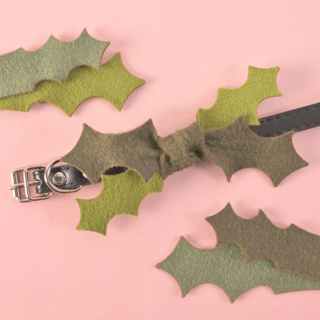 Handmade felt holly leaf dog collar bow on a black leather collar, surrounded by scattered felt holly leaves on a pink background