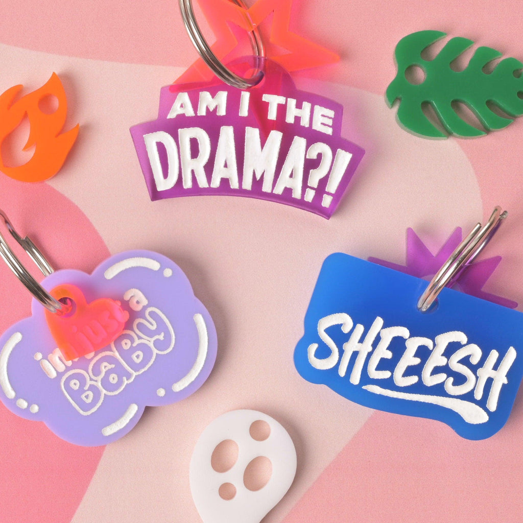 Flat lay of colorful acrylic pet ID tags with slang phrases—Am I the Drama, I’m just a Baby, Sheeesh—white engraved lettering on a pink background.