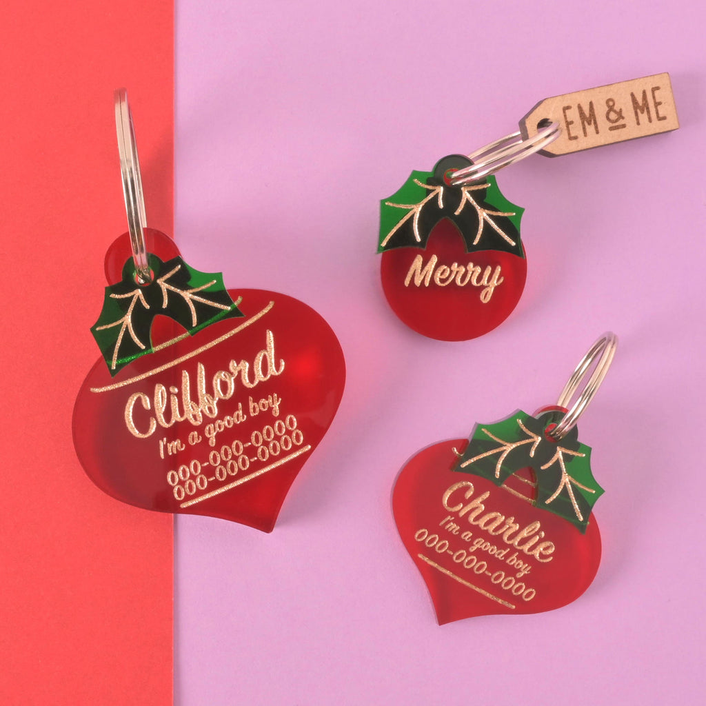 Set of red Christmas ornament-shaped acrylic dog tags with green holly detail and gold engraving – personalized for Clifford, Charlie, and Merry in multiple sizes