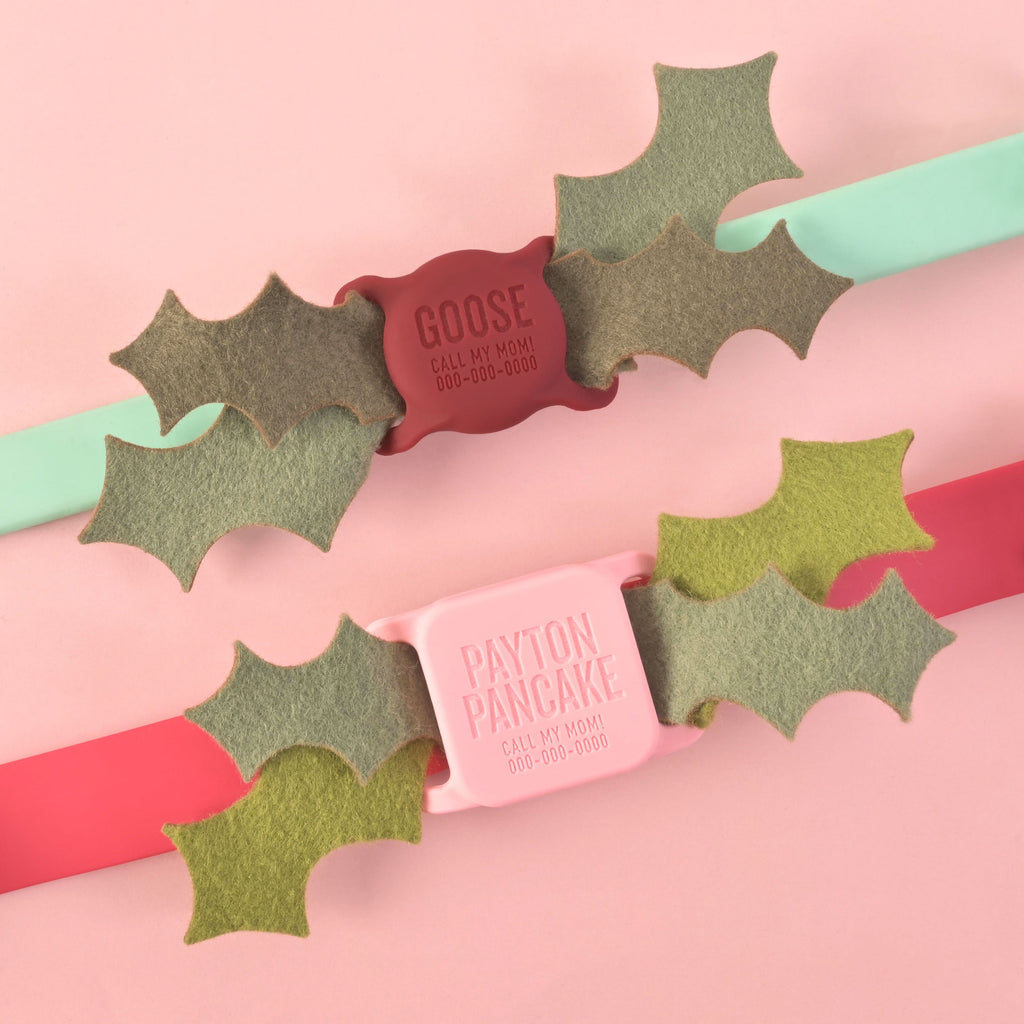 Two silicone pet ID tags on mint and red collars, each decorated with layered felt holly leaves. One tag reads 'GOOSE - CALL MY MOM!' in red, and the other reads 'PAYTON PANCAKE - CALL MY MOM!' in pink. Displayed on a soft pink background.