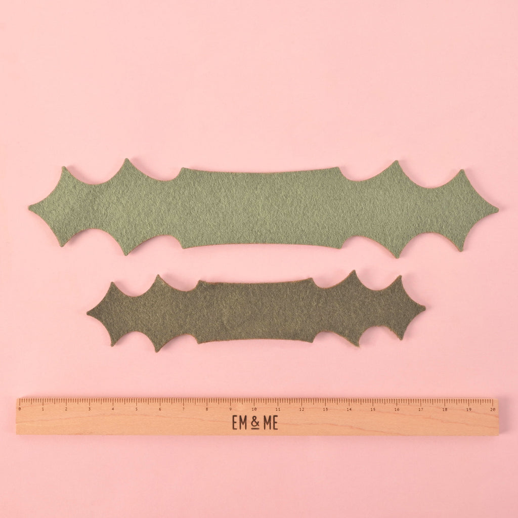 Two green felt holly leaf bow pieces in different sizes placed above a wooden EM & ME branded ruler on a pink background, showing size comparison for pet collar accessories.