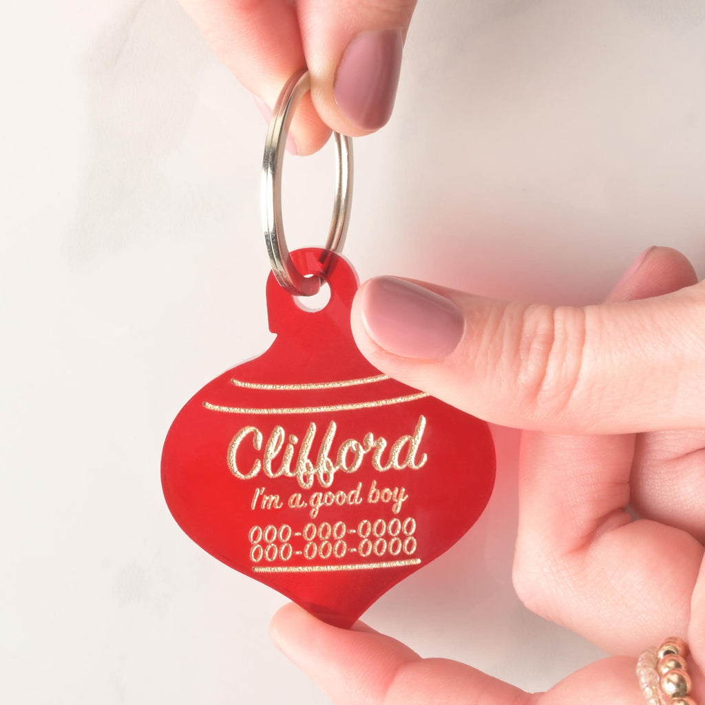 Hand holding red extra large Christmas ornament-shaped dog tag with gold engraving – personalized pet ID tag with custom name Clifford