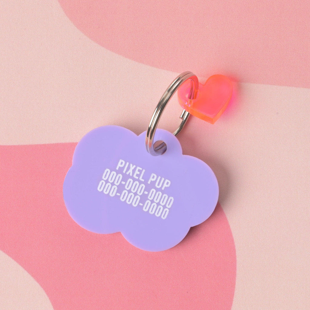 Lavender cloud acrylic pet ID tag back with “PIXEL PUP” and two phone numbers; split ring and neon pink heart charm on pink wavy background.