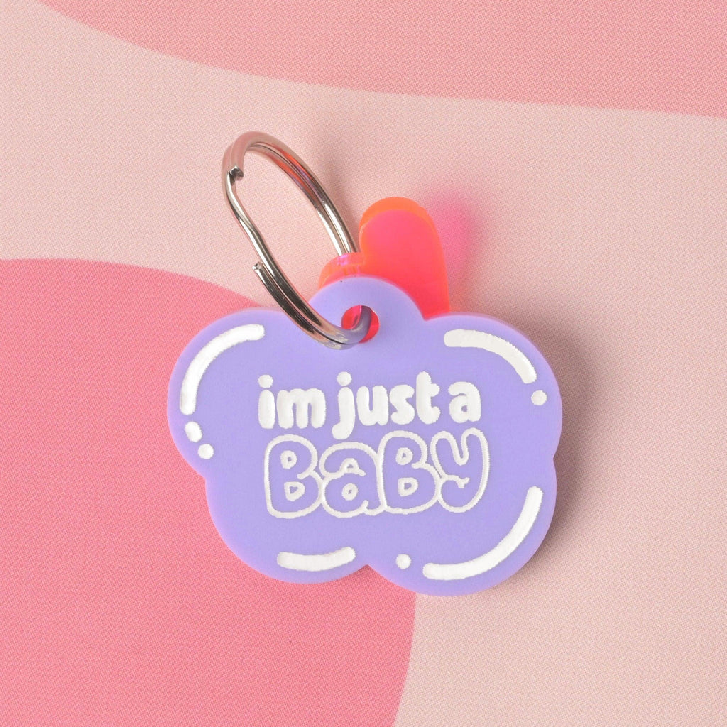 Lavender cloud-shaped acrylic pet ID tag reading “I’m just a baby,” on a stainless split ring with neon pink accent, styled on a pink wavy background.