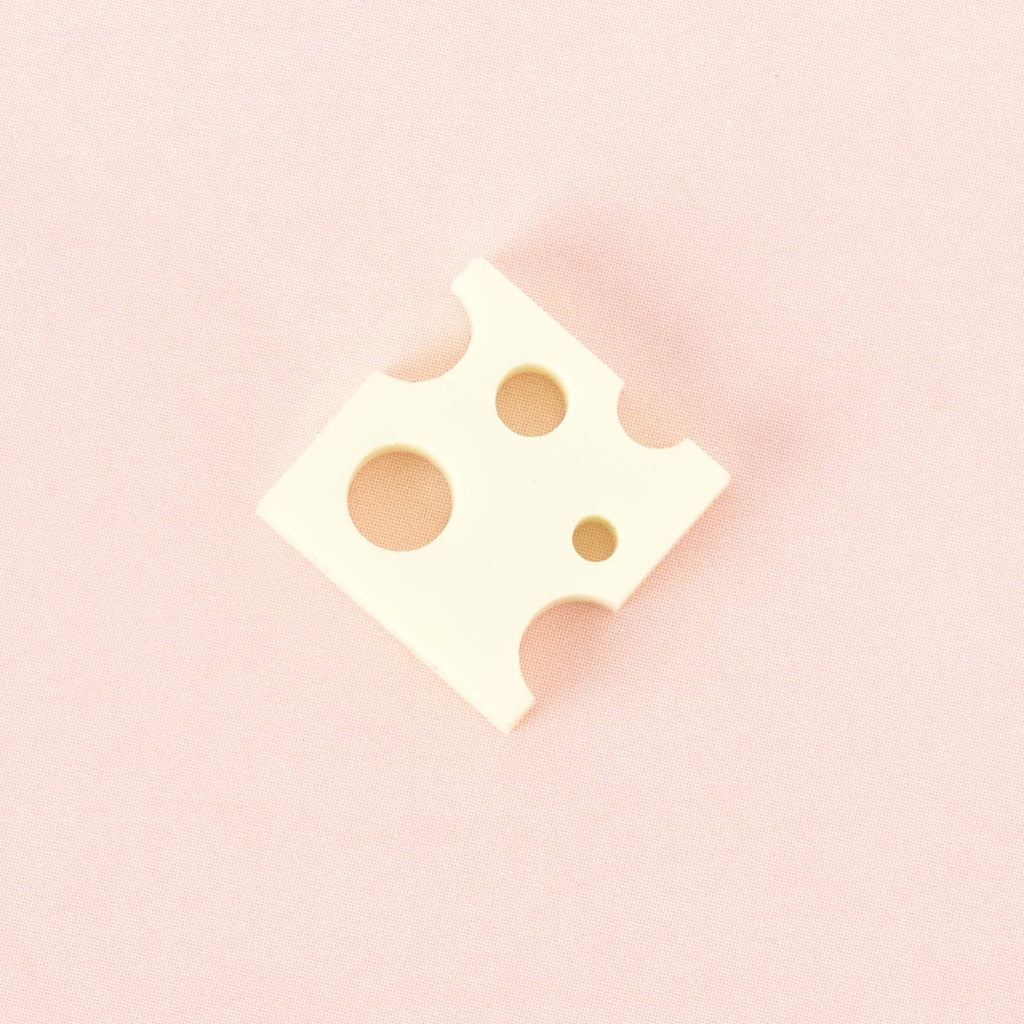 A single pale Yellow, cheese-shaped charm with various-sized holes. The item is shown on a solid pink background.