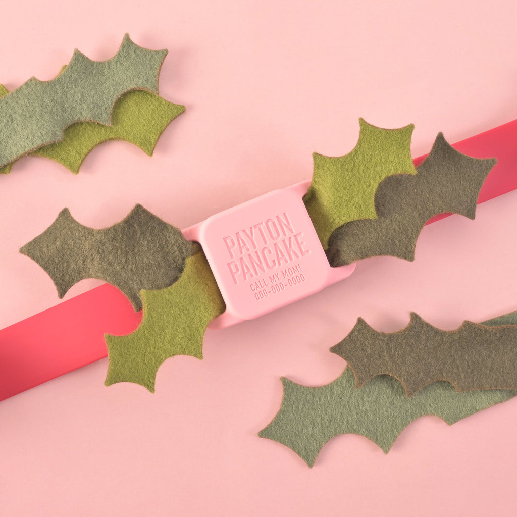 Pink square slide-on dog tag engraved with Payton Pancake, surrounded by felt holly leaves on a pastel pink background
