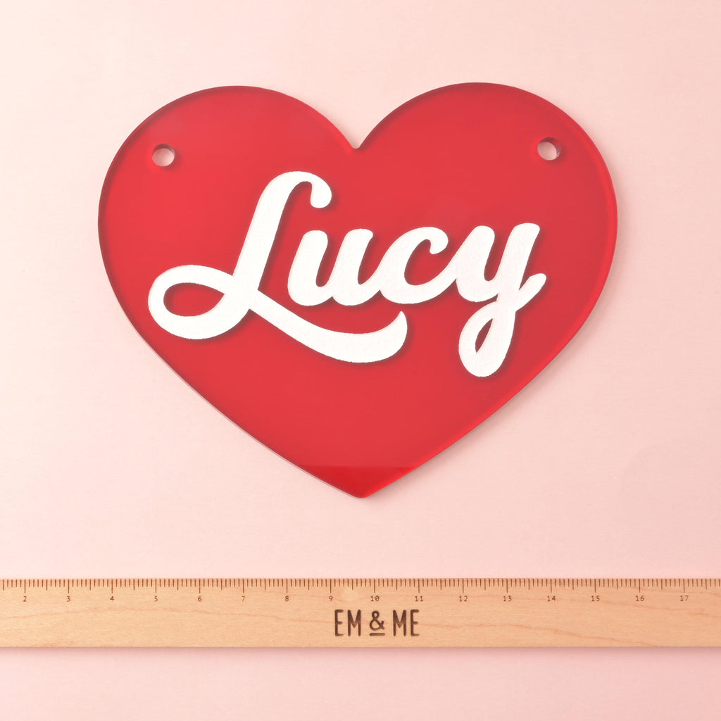 Red acrylic heart pet nameplate with two top holes and white script name “Lucy,” shown on a pink background with a wooden ruler for size reference.