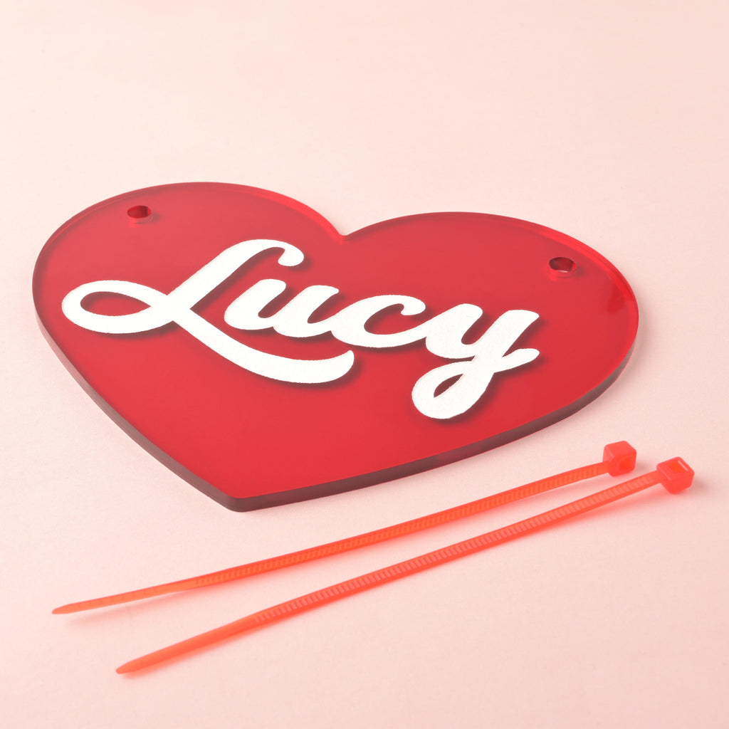 Angled close-up of red acrylic heart crate nameplate with two top holes and white script name “Lucy,” shown with two orange zip ties for attachment on a pink background.