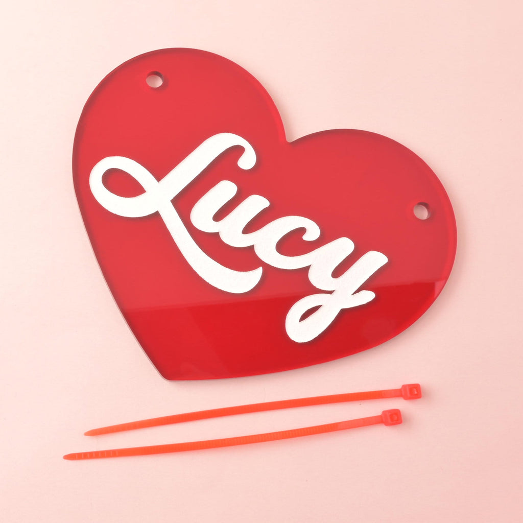 Red acrylic heart-shaped crate nameplate with white script name “Lucy,” two mounting holes, shown on pink background with two orange zip ties for attachment.