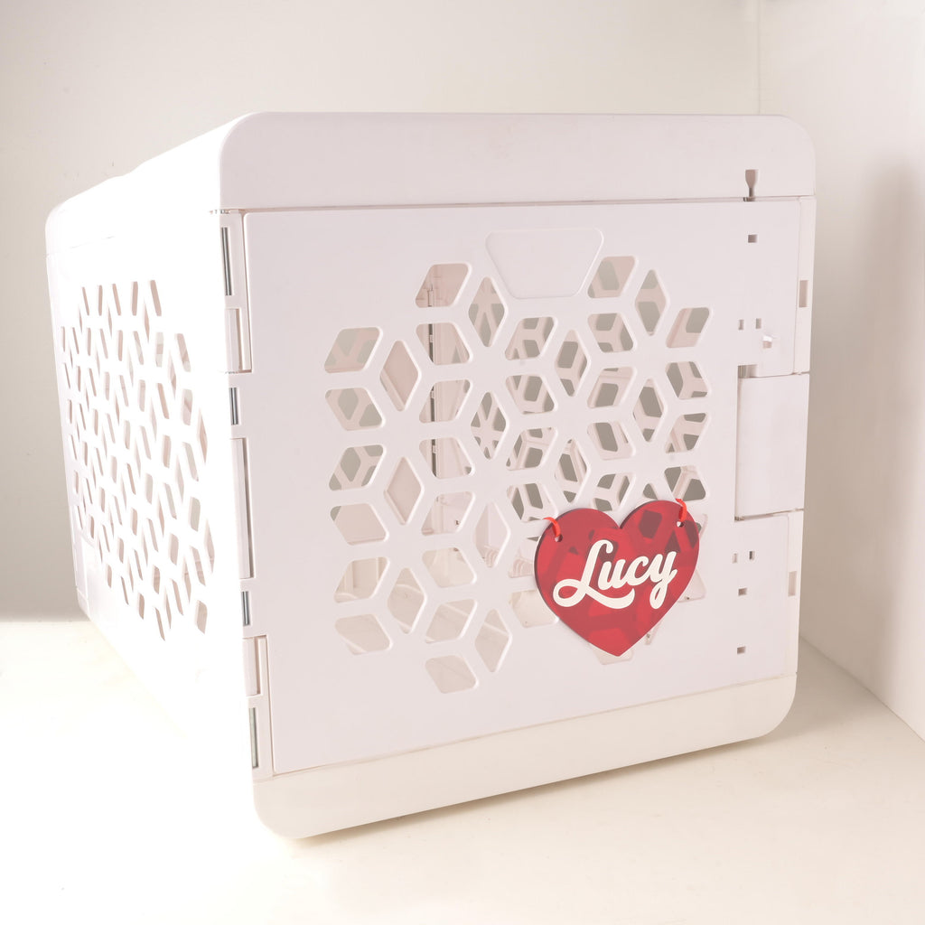 Red acrylic heart-shaped personalized crate nameplate with white script name “Lucy” attached to a modern white dog crate with honeycomb vents.