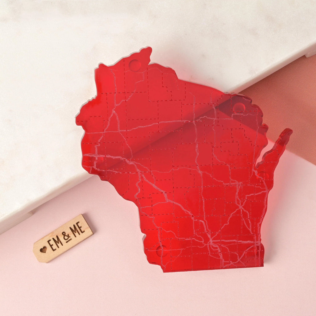Red acrylic Wisconsin-shaped coaster with laser-etched map detailing roads and counties, shown on soft pink and marble background with EM & ME tag.