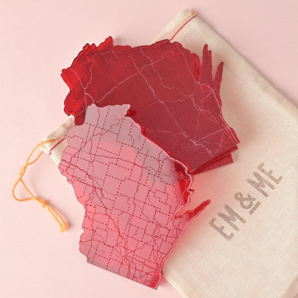 Set of red acrylic Wisconsin-shaped coasters with laser-etched roads and county grids, displayed on a cotton EM & ME drawstring bag against a pink background.